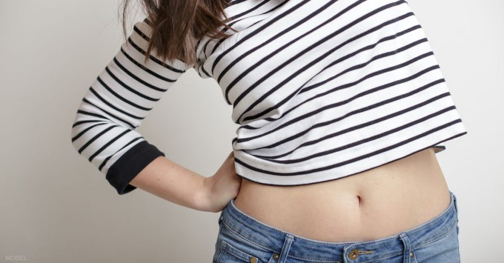 A woman (model) stretches, exposing her stomach on a dark white background